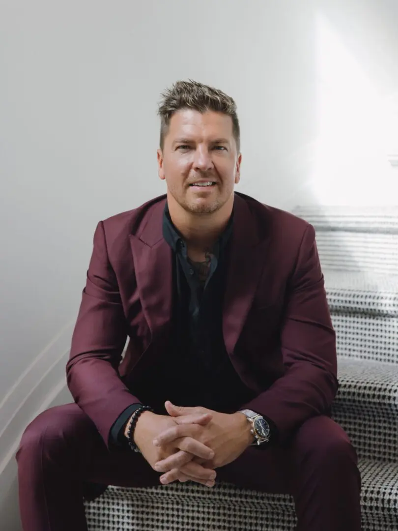 Man in a burgundy suit sitting on stairs, smiling.